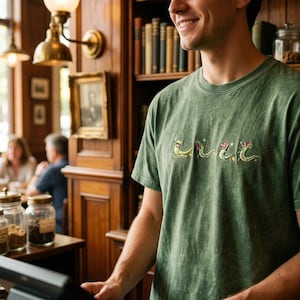 May include: A sage green mineral-washed t-shirt featuring an embroidered graphic of four small, light green lizards in various poses, accented with small white star shapes across the chest area of the garment.