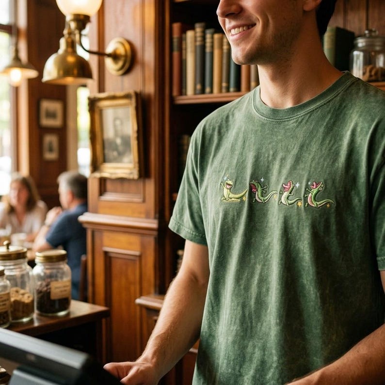May include: A sage green mineral-washed t-shirt featuring an embroidered graphic of four small, light green lizards in various poses, accented with small white star shapes across the chest area of the garment.
