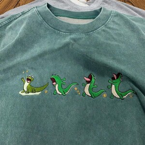 May include: A faded teal crewneck t-shirt featuring an embroidered graphic of four distinct green lizards in various poses. The lizards are depicted with open mouths and expressive faces, accented by small yellow embroidered stars scattered around them.