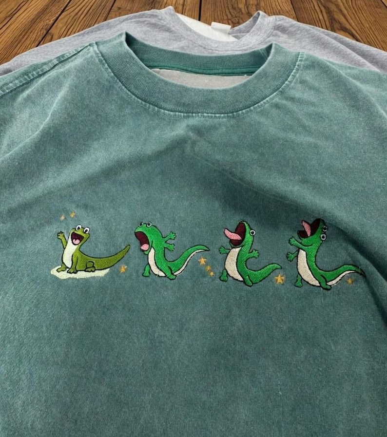 May include: A faded teal crewneck t-shirt featuring an embroidered graphic of four distinct green lizards in various poses. The lizards are depicted with open mouths and expressive faces, accented by small yellow embroidered stars scattered around them.