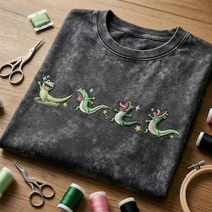May include: A charcoal grey acid-wash t-shirt featuring an embroidered design of four green lizards in various poses, each with an open mouth and tongue extended, accented by small embroidered yellow stars and light blue sparkles across the chest.