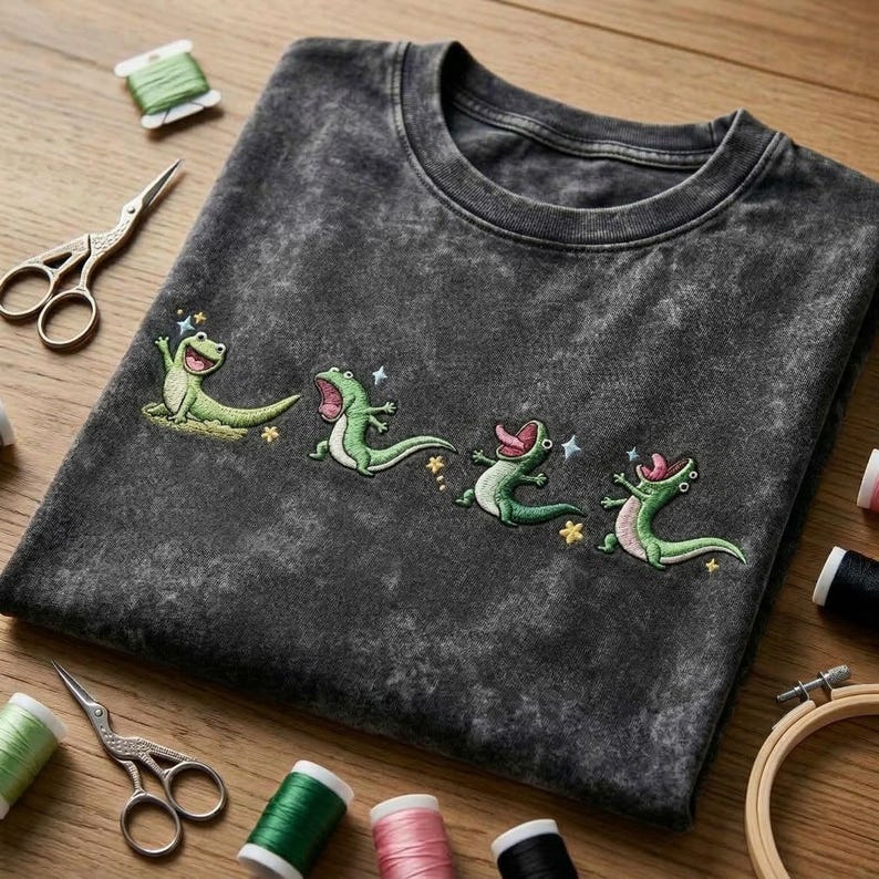 May include: A charcoal grey acid-wash t-shirt featuring an embroidered design of four green lizards in various poses, each with an open mouth and tongue extended, accented by small embroidered yellow stars and light blue sparkles across the chest.