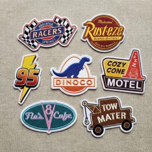 May include: A collection of embroidered patches featuring designs from the animated film Cars. Designs include "Radiator Racers," "Rusteze," "95," a blue dinosaur, "Dinoco," "Cozy Cone Motel," "Flo's Cafe," and "Tow Mater."