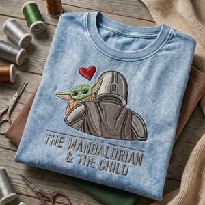 May include: A light blue acid-wash t-shirt featuring an embroidered graphic of The Mandalorian and The Child with a red heart above them. The text "THE MANDALORIAN & THE CHILD" is embroidered in dark gray thread below the character illustration.