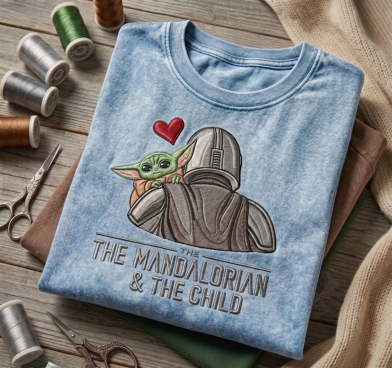May include: A light blue acid-wash t-shirt featuring an embroidered graphic of The Mandalorian and The Child with a red heart above them. The text "THE MANDALORIAN & THE CHILD" is embroidered in dark gray thread below the character illustration.