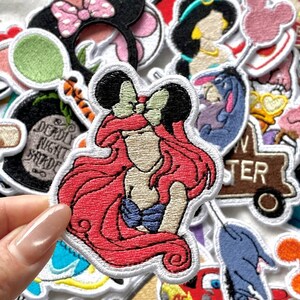 Może przedstawiać: Kolekcja kolorowych, haftowanych naszywek z r&oacute;żnymi postaciami Disneya. Naszywki przedstawiają Ariel z rudymi włosami, Minnie Mouse z r&oacute;żową kokardą i Jasmine. Naszywki mają białe obramowania i prawdopodobnie są przeznaczone na odzież lub akcesoria.