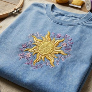 Op de afbeelding: Een lichtblauw T-shirt met ronde hals en een centraal borduurwerk van een goudgele zon, omringd door delicate paarse bloemranken en kleine roze bloemetjes. Het borduurwerk is voorzien van gestructureerde steken en gebogen zonnestralen.