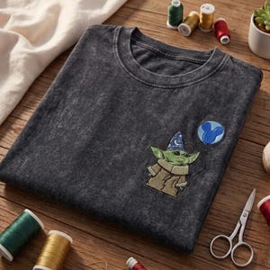 May include: A charcoal grey acid-wash t-shirt featuring an embroidered graphic of a green creature wearing a blue wizard hat with white stars, holding a blue balloon shaped like a mouse head. The shirt has a crew neckline and short sleeves.