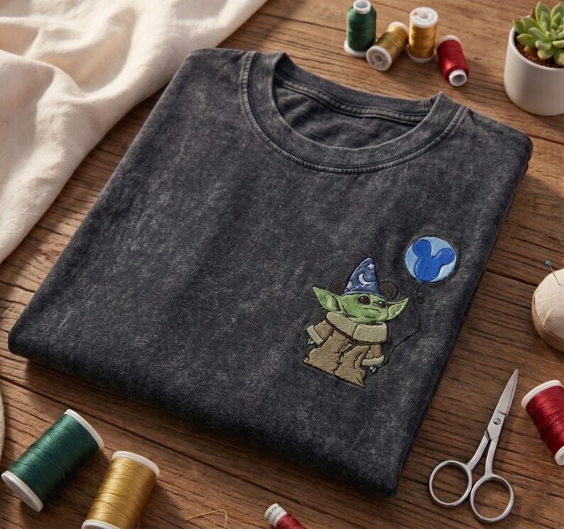 May include: A charcoal grey acid-wash t-shirt featuring an embroidered graphic of a green creature wearing a blue wizard hat with white stars, holding a blue balloon shaped like a mouse head. The shirt has a crew neckline and short sleeves.