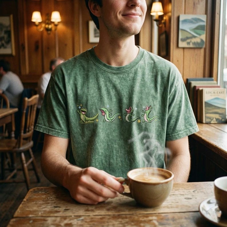 May include: A sage green mineral-washed t-shirt featuring a horizontal graphic print of four small, cartoonish green lizards in various poses, including one waving, one running, and two with open mouths, accented by small yellow star-shaped sparkles.