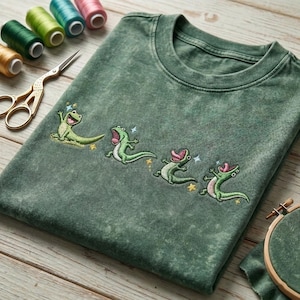May include: A mineral-washed forest green t-shirt featuring an embroidered design of four small, light green lizards in various playful poses, accented with tiny yellow and blue star-shaped details, arranged in a horizontal line across the chest.