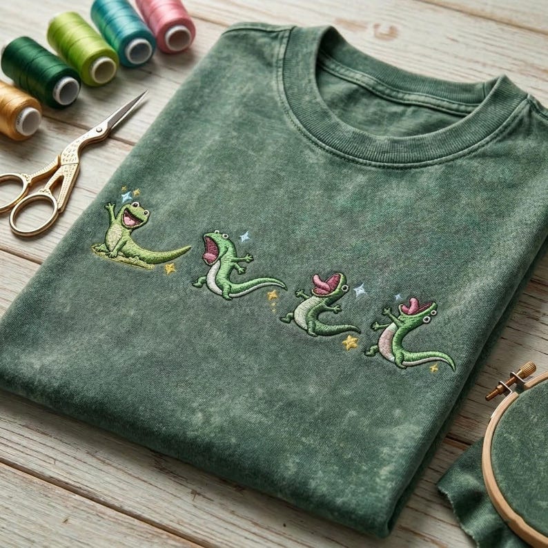 May include: A mineral-washed forest green t-shirt featuring an embroidered design of four small, light green lizards in various playful poses, accented with tiny yellow and blue star-shaped details, arranged in a horizontal line across the chest.