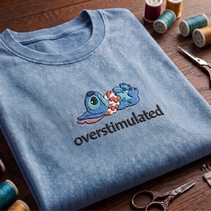 Pu&ograve; includere: Una t-shirt azzurro chiaro effetto lavaggio acido con il ricamo di un personaggio alieno blu sdraiato che indossa una collana di fiori rosa e bianca. Sotto il personaggio, la parola overstimulated &egrave; ricamata in lettere minuscole nere.