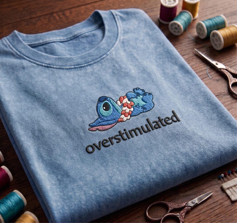 Pu&ograve; includere: Una t-shirt azzurro chiaro effetto lavaggio acido con il ricamo di un personaggio alieno blu sdraiato che indossa una collana di fiori rosa e bianca. Sotto il personaggio, la parola overstimulated &egrave; ricamata in lettere minuscole nere.
