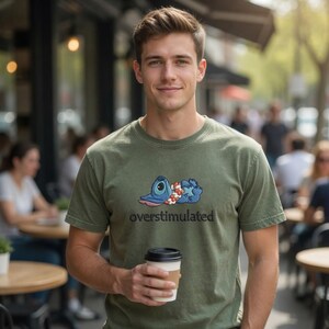 Pu&ograve; includere: Una t-shirt a maniche corte color verde oliva con la grafica di un personaggio alieno blu sdraiato con una collana di fiori al collo, posizionata sopra la scritta nera overstimulated, mentre si tiene in mano un bicchiere di caff&egrave; bianco.