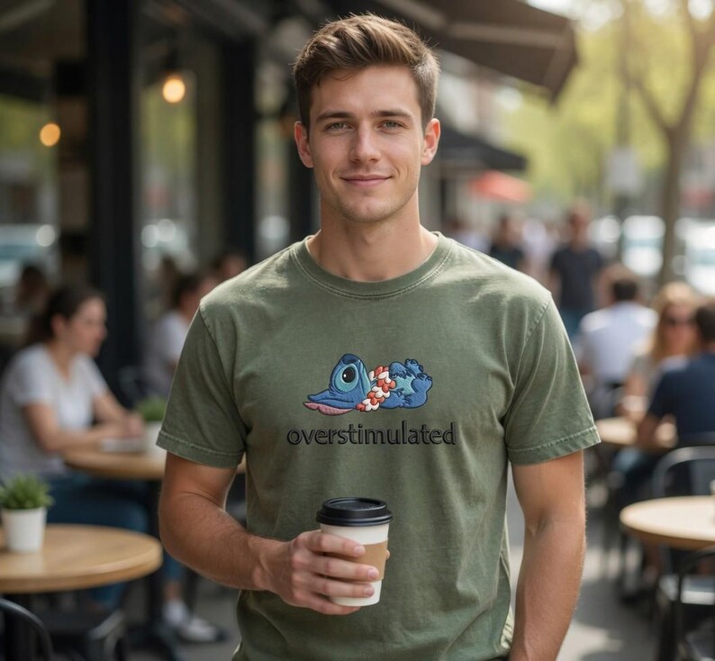 Pu&ograve; includere: Una t-shirt a maniche corte color verde oliva con la grafica di un personaggio alieno blu sdraiato con una collana di fiori al collo, posizionata sopra la scritta nera overstimulated, mentre si tiene in mano un bicchiere di caff&egrave; bianco.