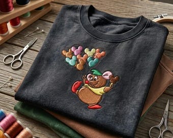 May include: A charcoal gray t-shirt featuring an embroidered graphic of a brown cartoon mouse wearing a green hat and yellow shirt, holding a cluster of colorful balloons shaped like mouse ears. The design includes pink, teal, and tan accents.