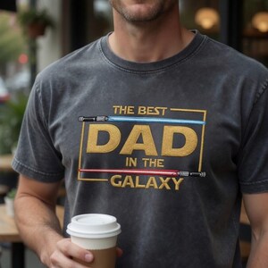 May include: A charcoal grey short-sleeve t-shirt featuring a gold embroidered rectangular frame containing the text THE BEST DAD IN THE GALAXY. A blue light saber is positioned above the word DAD and a red light saber is below the word IN THE.