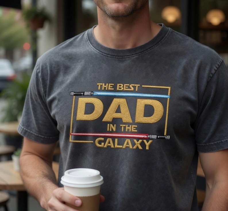 May include: A charcoal grey short-sleeve t-shirt featuring a gold embroidered rectangular frame containing the text THE BEST DAD IN THE GALAXY. A blue light saber is positioned above the word DAD and a red light saber is below the word IN THE.