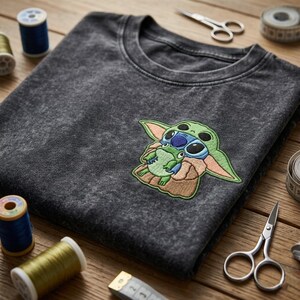 May include: A charcoal gray acid-wash t-shirt featuring an embroidered patch of a green alien character holding a small green frog. The shirt is surrounded by sewing supplies including blue and gold thread spools, silver scissors, and a measuring tape.