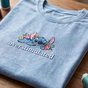 K&ouml;nnte beinhalten: Ein hellblaues T-Shirt mit Rundhalsausschnitt und einer Stickerei, die eine blaue au&szlig;erirdische Figur zeigt, welche von kleinen rosa, gelben und t&uuml;rkisfarbenen Blumen umgeben ist, mit dem Wort "overstimulated" in wei&szlig;em Garn darunter.