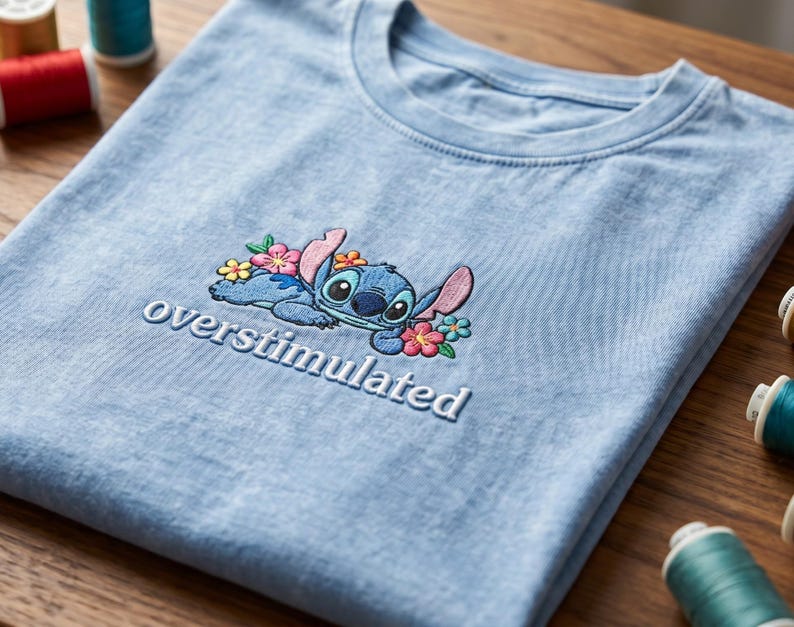 K&ouml;nnte beinhalten: Ein hellblaues T-Shirt mit Rundhalsausschnitt und einer Stickerei, die eine blaue au&szlig;erirdische Figur zeigt, welche von kleinen rosa, gelben und t&uuml;rkisfarbenen Blumen umgeben ist, mit dem Wort "overstimulated" in wei&szlig;em Garn darunter.