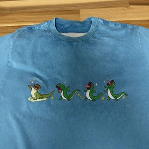 May include: A light blue crew neck t-shirt featuring an embroidered graphic of four green lizards in various poses. The lizards are depicted with open mouths and tongues, surrounded by small yellow stars and white sparkles across the chest area.