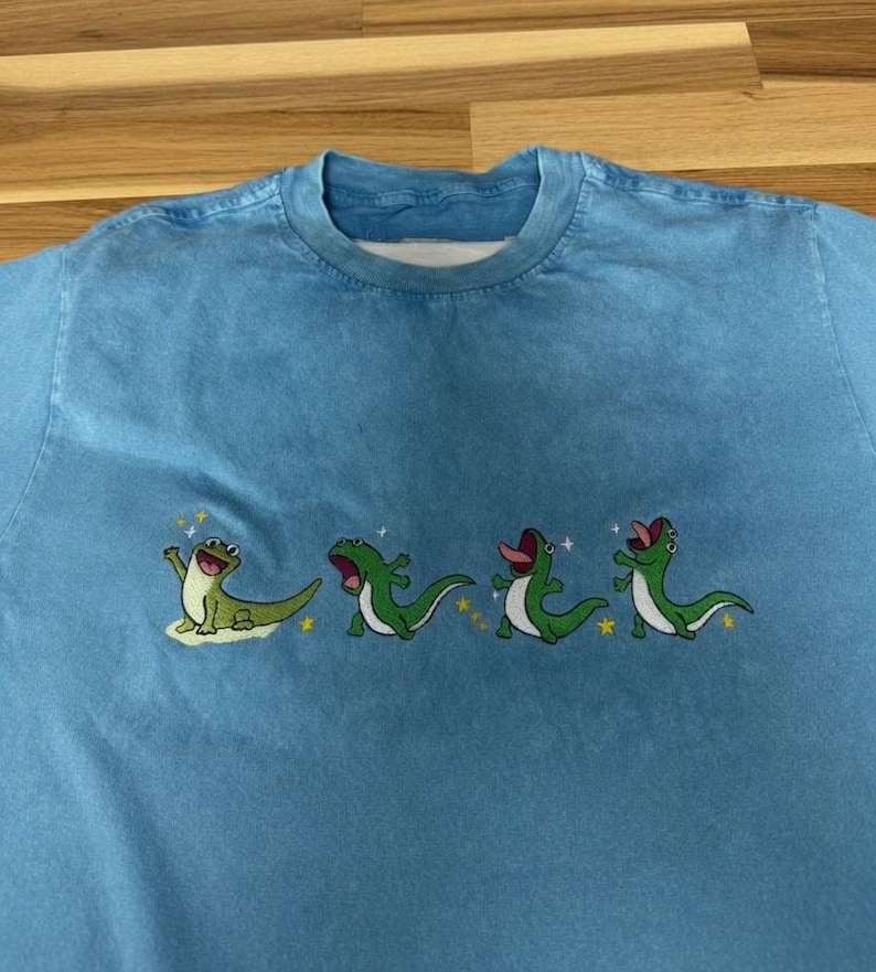 May include: A light blue crew neck t-shirt featuring an embroidered graphic of four green lizards in various poses. The lizards are depicted with open mouths and tongues, surrounded by small yellow stars and white sparkles across the chest area.