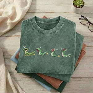 May include: A dark green crew neck t-shirt featuring a graphic print of four small, light green lizards in various poses, accented with tiny yellow stars and white sparkles. The fabric has a subtle, washed-out, mottled texture throughout.