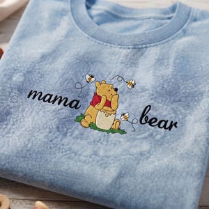 May include: A light blue tie-dye t-shirt featuring an embroidered graphic of a yellow bear wearing a red shirt, sitting on green grass with a honey pot. The text mama bear is embroidered in black script on either side of the bear with small bees.