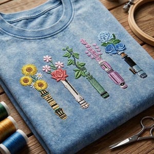 K&ouml;nnte beinhalten: Ein hellblaues T-Shirt in Acid-Wash-Optik mit f&uuml;nf Stickereien, die metallische Griffe zeigen, welche mit verschiedenen Blumen wie Sonnenblumen, einer roten Rose, gr&uuml;nen Ranken, Lavendel und blauen Hortensien verziert sind.