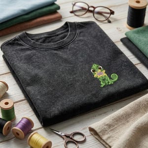 May include: A charcoal gray mineral-washed t-shirt featuring a small embroidered green chameleon holding a yellow and purple flower on the left chest. The shirt is folded and surrounded by sewing supplies including thread spools and scissors.