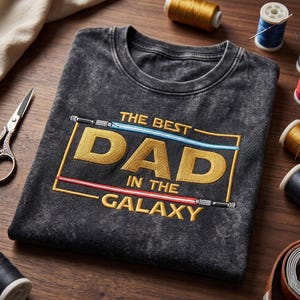 May include: A charcoal grey acid-wash t-shirt featuring an embroidered design with the text THE BEST DAD IN THE GALAXY in gold letters. The text is framed by a blue light saber at the top and a red light saber at the bottom of the rectangular border.
