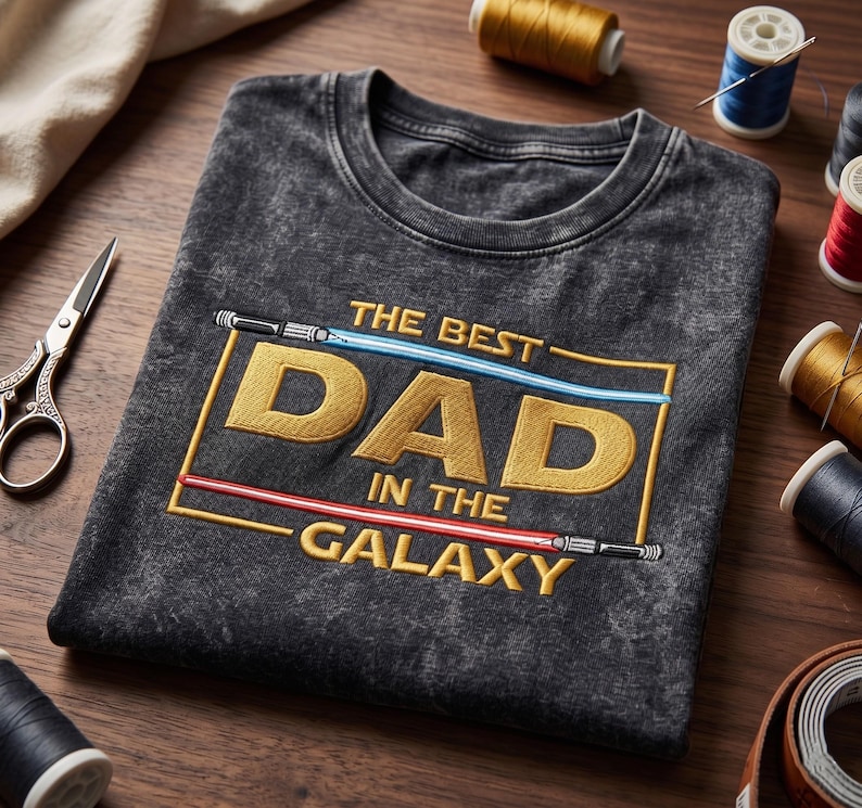 May include: A charcoal grey acid-wash t-shirt featuring an embroidered design with the text THE BEST DAD IN THE GALAXY in gold letters. The text is framed by a blue light saber at the top and a red light saber at the bottom of the rectangular border.