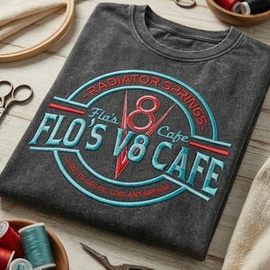 May include: A charcoal gray t-shirt featuring an embroidered logo for Flo's V8 Cafe in Radiator Springs. The design includes red and light blue stitching with the text Flo's V8 Cafe, Radiator Springs, and Lugnuts-Gas-Oil-Coolant-Grease.