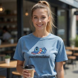 Pu&ograve; includere: Una t-shirt a maniche corte azzurra con un ricamo raffigurante un personaggio alieno blu che indossa una collana di fiori, sdraiato. Sotto il disegno, la parola "overstimulated" &egrave; ricamata in lettere minuscole nere sul petto.