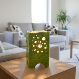 Może przedstawiać: Zielona ceramiczna lampa o prostokątnym kształcie i falistym górnym brzegu. Lampa ma liczne okrągłe wycięcia o różnych rozmiarach, przez które prześwituje światło. Stoi na drewnianym stole w salonie.