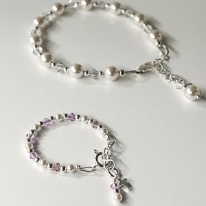 Puede incluir: Dos pulseras de plata con perlas blancas y detalles de cristal. La pulsera más pequeña tiene un pequeño dije de cruz de plata y cristales morados.
