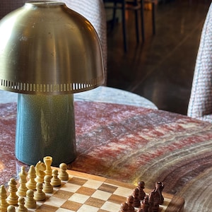 Puede incluir: Un tablero de ajedrez de madera con un patrón de tablero de ajedrez marrón claro y marrón oscuro. El tablero de ajedrez está configurado con todas las piezas en sus posiciones de inicio. El tablero de ajedrez está sobre una mesa con una lámpara dorada en el fondo.