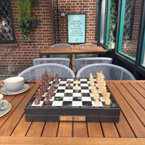 May include: A chess set with a black and white marble-effect board and wooden pieces. The set is in a dark wooden box with a gold nameplate. The chess pieces are brown and cream. The set is on a wooden table.