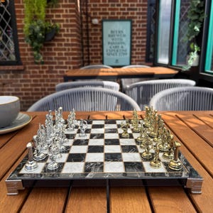 Könnte beinhalten: Ein Schachspiel mit einem schwarz-weiß marmorierten Schachbrett und silbernen und goldenen Schachfiguren. Die Schachfiguren sind auf dem Brett angeordnet und bereit für ein Spiel. Das Brett steht auf einem Holztisch.