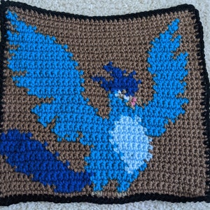 May include: Crocheted square featuring a blue and brown bird design. The bird has a blue body, brown wings, and a blue head with a white beak.