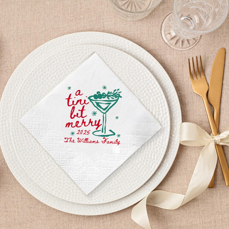Op de afbeelding: Witte vierkante servet met een rood en groen cocktailontwerp en de tekst "a tini bit merry *2025* The Williams Family". De servet ligt op een wit bord met gouden bestek en een cr&egrave;mekleurig lint.
