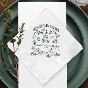 May include: White paper napkin with green text and illustrations. The text reads "Mountain Views Before I Do's" with illustrations of a mountain range, trees, and camping gear. The napkin is on a white card and a green plate.