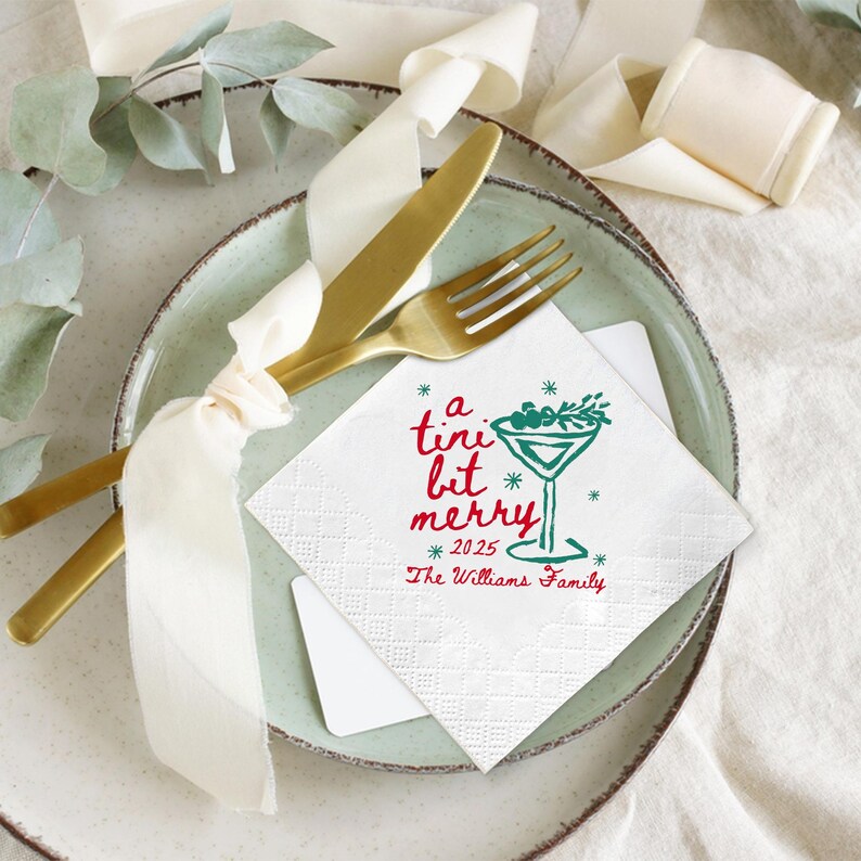 Op de afbeelding: Een feestelijke tafelsetting met een gouden vork en mes, vastgebonden met een cr&egrave;mekleurig lint. Een witte servet met de tekst "a tini bit merry" en een martini glas ontwerp ligt op een groene bord. Het jaar 2025 staat ook op de servet.