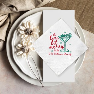 Op de afbeelding: Witte cocktailservet met de tekst "a tini bit merry" in rood en groen, en een martini glas illustratie. Het jaar 2025 en "The Williams Family" staan ook op de servet. De servet ligt op een wit bord met decoratieve bloemen.