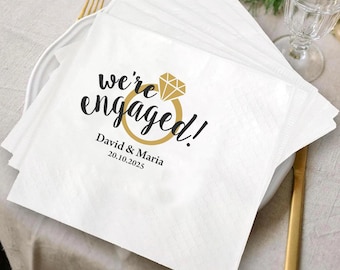 May include: White paper napkins stacked on a white plate. The napkins have the text "we're engaged!" in black script with a gold ring graphic. Below the text is "David & Maria 20.10.2025".
