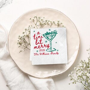Op de afbeelding: Een witte cocktailservet met de tekst "a tini bit merry 2025" in rood en groen. Een groene martini glas afbeelding is ook te zien. De servet ligt op een cr&egrave;mekleurige schotel, versierd met witte bloemen.
