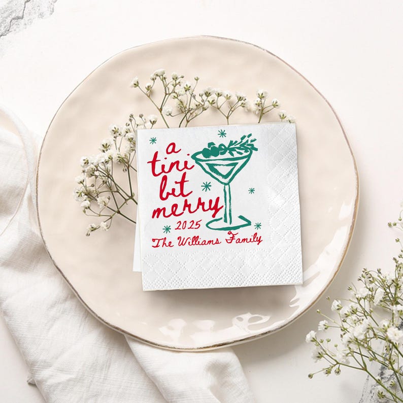 Op de afbeelding: Een witte cocktailservet met de tekst "a tini bit merry 2025" in rood en groen. Een groene martini glas afbeelding is ook te zien. De servet ligt op een cr&egrave;mekleurige schotel, versierd met witte bloemen.