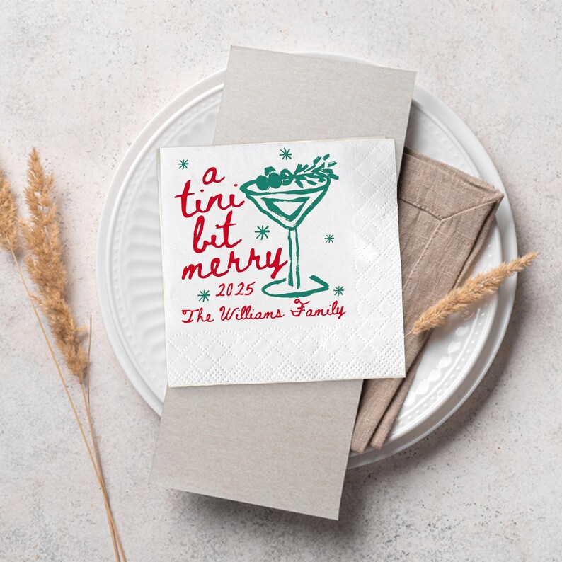 Op de afbeelding: Witte cocktailservet met een groen martini glas ontwerp en de woorden "a tiny bit merry" in het rood. Het jaar 2025 en "The Williams Family" staan ook op de servet. De servet ligt op een wit bord.